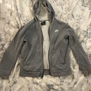 Grå hoodie från Nike med dragkedja - Grå hoodie från Nike med vit logga på bröstet. Tröjan har luva, dragkedja framtill och två fickor. Mjuk insida och långärmad modell, i lappen på tröjan står det storlek M men den är snarare Xs. 