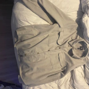 Beige overshirt från H&M i storlek L - Snygg beige overshirt från H&M i storlek L passar även dom som har storlek M. Skjortan har två stora bröstfickor med lock, klassisk krage och knäppning med beigea knappar framtill. Materialet är mjukt och känns som bomull, perfekt för lager-på-lager. Enkel och stilren design.