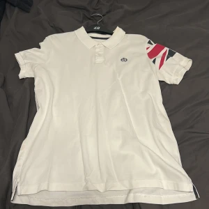 Vit piké från Henri Lloyd med tryck - Vit pikétröja från Henri Lloyd i bomull, regular fit. Klassisk krage med knappar och coolt tryck i rött, vitt och blått på ena ärmen. Liten broderad logga på bröstet. Perfekt för dig som gillar stilren design med en twist.