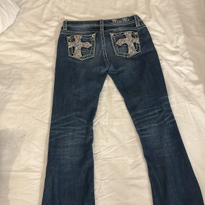 Bootcut jeans från Miss Me med broderade fickor - Snygga mörkblå bootcut jeans från Miss Me med kontrastsömmar och broderade bakfickor med korsdetaljer. Jeansen har låg midja och klassisk femficksdesign. Materialet är slitstarkt denim och passformen är skinny med utsvängda ben (bootcut)