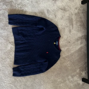 Marinblå stickad tröja från Polo Ralph Lauren - Säljer en marinblå stickad tröja från Polo Ralph Lauren med klassiskt kabelstickat mönster och röd broderad logga på bröstet. Tröjan har rund hals och långa ärmar