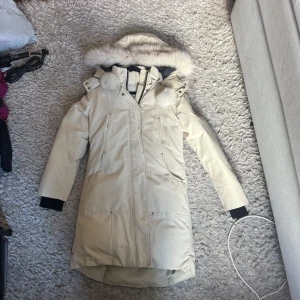 Beige dunjacka Moose Knuckles - Snygg beige dunjacka från Moose Knuckles med pälskantad huva och broderad hockeypatch på insidan. Jackan har långa ärmar, dragkedja och knappar framtill samt svarta muddar vid ärmslut. Perfekt för kalla vinterdagar.