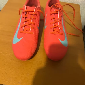 Nike fotbollsskor i neonrosa och turkos - Snygga fotbollsskor från Nike i neonrosa med turkosa swoosh-loggor och detaljer. Skorna har snörning och en låg profil, perfekt för snabba rörelser på planen. Ovandelen är i syntetmaterial för lätt känsla och bra bollkontroll. Riktigt coola färger som sticker ut på matchen.