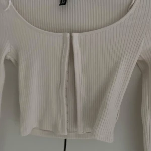 Ljusbeige ribbad croppad topp - Säljer en ljusbeige croppad topp med långa ärmar och ribbad struktur. Toppen har en djup rundad urringning och knäppning framtill. Perfekt för att styla med höga jeans eller kjol.