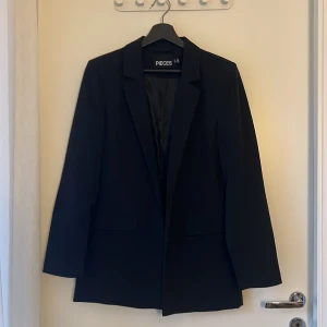 Svart kavaj från Pieces, storlek M - Stilren svart kavaj från Pieces i klassisk modell med krage. Kavajen är helfodrad och har en rak passform som passar till många olika looks. Perfekt för dig som gillar en clean och tidlös stil.
