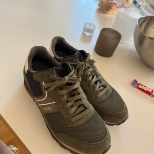 Olivgröna sneakers från BOSS - Snygga sneakers från BOSS i olivgrönt med svarta och vita detaljer. Skorna har grov sula, snörning och är tillverkade i en mix av mocka och textil. Sidan pryds av BOSS-logga och silverfärgade ränder för en sportig look.