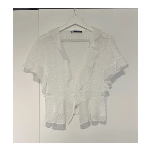 Vit volangblus från Zara - Supersöt vit blus från Zara med prickigt mönster, volangdetaljer längs ärmar och framkant samt v-ringning. Blusen har korta ärmar och knappar framtill. Använd få gånger.