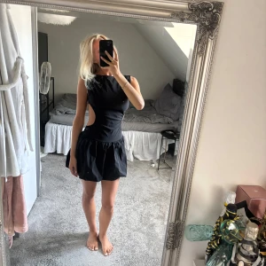 Svart kort klänning med öppen rygg - Supersnygg svart kort klänning med markerad midja och voluminös kjol. Klänningen har ärmlös design och cut-outs i sidorna som ger en öppen rygg. 