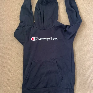Svart Champion hoodie - Svart hoodie från Champion i storlek XL med klassisk logga broderad på bröstet. Tröjan har huva, magficka och långa ärmar. Perfekt för en chill och sportig stil. Materialet är mjuk bomullsmix som känns skön mot huden.