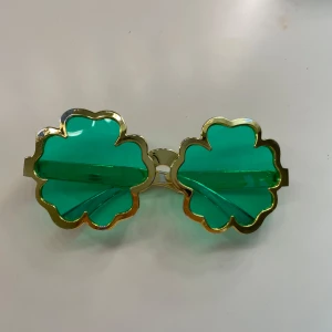 Gröna klöverformade solglasögon - Säljer ett par unika solglasögon med klöverformade gröna linser och guldfärgade kanter. Perfekta för dig som vill sticka ut och addera lite extra färg till din stil. Ramen är i plast och har en cool, lekfull design.