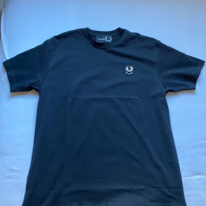 Svart oversized t-shirt Raf Simons - Svart oversized t-shirt från Raf Simons x Fred Perry med rund hals och korta ärmar. På bröstet sitter en broderad patch med Fred Perry-kransen och Raf Simons-loggan. Tillverkad i mjuk bomull, perfekt för en clean och stilren look.                                         Tröjan är HELT ny och aldrig använd med etiketten kvar.    Nypris på tröjan är 1600kr och säljer den för halva priset!!! 🔥🔥
