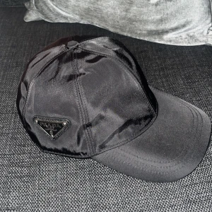 Svart keps från Prada - Snygg svart keps från Prada i glansig nylon med klassisk böjd skärm och justerbar rem bak. Kepsen har Pradas ikoniska metallogo på sidan och är tillverkad i återvunnen nylon. Perfekt accessoar för en clean och lyxig look. Pris kan diskuteras 