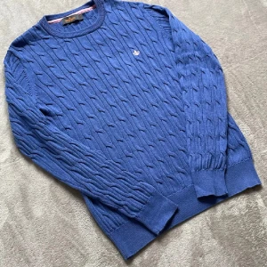 Blå kabelstickad tröja Morris M - Snygg blå kabelstickad tröja från Morris i storlek M. Tröjan har ett klassiskt mönster och en rosa broderad logga på bröstet. Rund hals och ribbade muddar vid ärmslut och nederkant. Perfekt för dig som gillar stilrena och tidlösa plagg. Säljer för den är för stor för mig har själv aldrig använt den