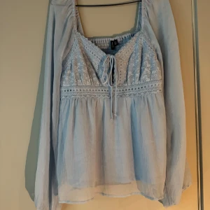 Ljusblå blus med spets och knyt - Supersöt ljusblå blus från Vero Moda med broderade spetsdetaljer och knyt framtill. Blusen har långa, luftiga ärmar och smockad rygg för extra komfort. 🥰