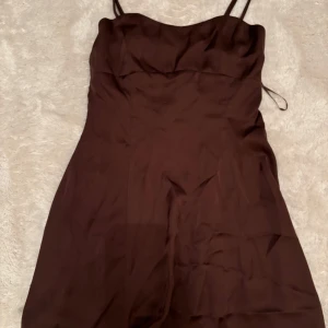 Brun kort klänning från Gina Tricot - Säljer en brun kort klänning från Gina Tricot i storlek XS. Klänningen har smala axelband och en figurnära passform med lätt utställd kjol. Materialet är mjukt och har en lätt glansig finish, perfekt för att skapa en chic look. Baksidan har smock för extra komfort. Nytt skick
