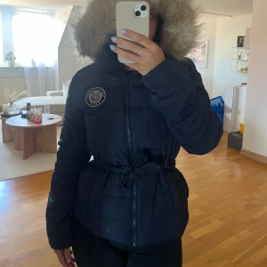 Svart pufferjacka med fuskpäls från Svea - Säljer en svart pufferjacka från Svea med stor huva och fuskpäls. Jackan har dragkedja, knytband i midjan och en snygg logga på bröstet. Tillverkad i polyester och perfekt för kalla dagar. Fodret är rosa/röd för en extra pop av färg.