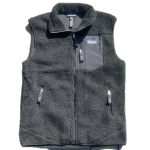 Svart fleeceväst från Patagonia - Mysig svart fleeceväst från Patagonia med hög krage och hel dragkedja framtill. Västen har tre fickor med dragkedja, varav en bröstficka med logga. Perfekt för lager-på-lager och outdoor-vibe.