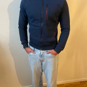 Marinblå zip hoodie Helly Hansen M - Snygg marinblå hoodie från Helly Hansen med hel dragkedja och röda detaljer vid fickorna och bröstet. Tröjan har lång ärm, ribbade muddar och HH-logga på ärmen. Perfekt för dig som gillar sportig och clean stil.