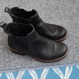 Svarta chelsea boots från Ecco - Säljer ett par svarta chelsea boots från Ecco med grov sula och rund tå. Skorna är tillverkade i robust skinn med elastiska paneler på sidorna och dragflikar bak för enkel påtagning. Perfekt för dig som gillar klassisk stil med edge.