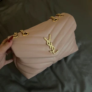 Rosa ysl väska  - Säljer en lyxig beige axelväska från Saint Laurent med ikonisk YSL-logga i guld framtill. Väskan är quiltad och har guldfärgade kedjedetaljer samt magnetlås. Insidan har dragkedjefack och flera fack för smart organisering. Perfekt för dig som vill ha en snygg och stilren väska. Fråga om fler bilder 