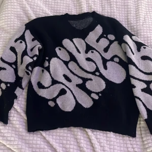 Svart stickad tröja  - Cool svart stickad tröja med unikt mönster i ljusgrått som slingrar sig över hela tröjan. Tröjan har rund halsringning och långa ärmar. Perfekt för dig som vill sticka ut med en grafisk och edgy stil.