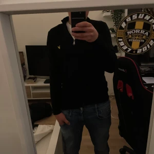 Svart half zip tröja från Lyle & Scott - Svart långärmad tröja från Lyle & Scott med half zip och klassisk gul logga på bröstet. Tröjan har en hög krage och är i mjukt material, perfekt för lager-på-lager. Passformen är normal och stilen är clean och enkel.
