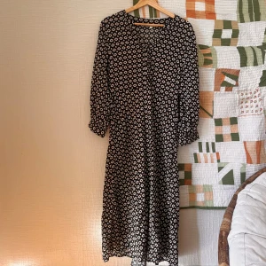 Svart långklänning med mönster från Indiska - Långklänning från Indiska i svart med vitt geometriskt mönster. Klänningen har långa ärmar med resår vid ärmslut, rundad hals och knappar framtill. Materialet är mjukt och lätt, perfekt för dig som gillar boho-vibe och snygga prints.