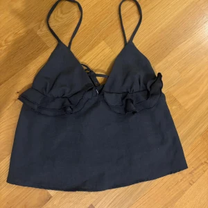 Svart linnetopp  - Snygg svart linnetopp med tunna axelband, volangdetaljer över bysten och snörning både fram och bak, storlek S. Toppen har en croppad passform och är perfekt för varma dagar. 