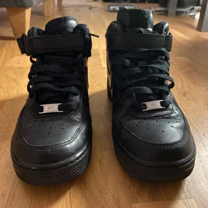 Nike Air Force 1 Mid Svarta Sneakers - Svarta Nike Air Force 1 Mid sneakers i skinn med klassisk snörning, kardborreband vid ankeln och perforerade detaljer på tån. Swoosh-logga på sidan och AIR-detalj på sulan. Perfekta för dig som gillar en clean och tidlös look.