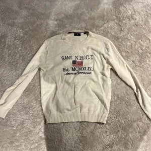 Vit stickad tröja från GANT - Vit stickad tröja från GANT med tryck på bröstet och amerikansk flagga. Rund hals med ribbad kant och ribbade muddar vid ärmslut och nederkant. Snygg och klassisk design, perfekt för dig som gillar preppy stil.