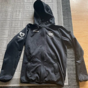 Svart vindjacka CCM med huva - Svart vindjacka från CCM med dragkedja, huva med snörning och två fickor framtill. Jackan har tryck med Hofors Hockey Camp på bröstet och Hofors-logga på ärmen. Perfekt för dig som gillar sportig stil och hockey.