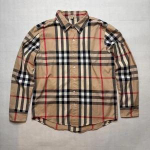 Rutig skjorta från Burberry Brit M - Klassisk rutig skjorta från Burberry Brit i beige, svart, rött och vitt. Skjortan har lång ärm, knäppning framtill och det ikoniska rutmönstret. Tillverkad i 100% bomull för en skön, stilren och tidlös känsla.  Storlek: Märkt som M, men passar även en S. Skick: Mycket fint, finns en liten smutsfläck på vänster ärm/hand som troligen går bort i tvätt. Äkthet: 100% autentisk Burberry.  📦 Skickas inom 1–3 dagar. 📩 För mer information eller frågor, kontakta mig gärna.