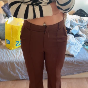 Bruna kostymbyxor med hög midja - Snygga bruna kostymbyxor med hög midja och raka ben. Byxorna har diskreta fickor i sidorna och stängs med dragkedja och knapp framtill. Perfekta för en clean och stilren look. Materialet känns mjukt och följsamt.