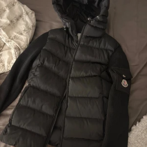 Svart Moncler pufferjacka med huva - Svart pufferjacka från Moncler med huva och dragkedja. Jackan har quiltad kropp i blankt material och ribbstickade ärmar med ficka och Moncler-logga på ena armen. Perfekt för kalla dagar och riktigt snygg streetstil. Obs ej äkta