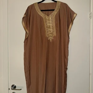 Brun kaftanklänning med guldiga broderier - Säljer en lång brun kaftanklänning med vackra guldiga broderier runt halsringningen och ärmsluten. Klänningen har lös passform och är ärmlös med en elegant v-ringning. Perfekt för dig som gillar unika detaljer och bekväma plagg.