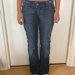 Levis lågmidjade blå bootcut jeans - Säljer ett par Levisjeans i klassisk blå tvätt med bootcut passform och låg midja. Jeansen har snygga detaljer som kontrastsömmar, fem fickor och Levis patch baktill. Materialet är slitstarkt denim i bomull. Perfekt för dig som gillar retrovibe och cool stil.