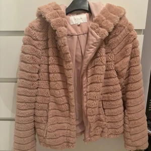 Rosa teddyjacka från Vila - Supermjuk teddyjacka från Vila i rosa, med fluffig struktur och horisontella ränder. Jackan har huva, dragkedja + knappar framtill och är helfodrad i matchande färg. Storlek S. OBS ena fickan är lite trasig. 