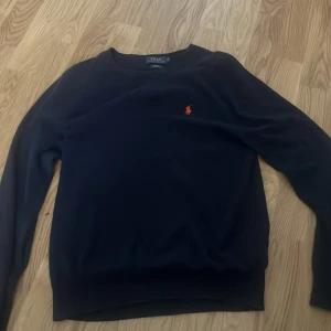Mörkblå tröja från Polo Ralph Lauren - Snygg mörkblå tröja från Polo Ralph Lauren i slim fit med klassisk orange broderad logga på bröstet. Tillverkad i mjuk pima bomull, perfekt för dig som gillar stilrena och tidlösa plagg. Rund hals och långa ärmar ger en clean look.