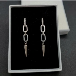 Set med silverfärgade örhängen och halsband - Säljer ett set med coola silverfärgade örhängen med kedjelänkar och spetsiga hängen samt ett matchande halsband med en kedja och en hängande vit pärldroppe formad som en ängel. Perfekt för dig som gillar stilrena och trendiga accessoarer.