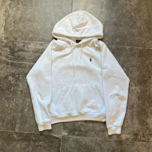 Vit hoodie från Polo Ralph Lauren - Klassisk vit hoodie från Polo Ralph Lauren med broderad marinblå logga på bröstet. Tröjan har huva med snörning, känguruficka och ribbade muddar. Tillverkad i mjuk bomullsblandning med polyester för extra komfort. Perfekt för en clean och stilren look.