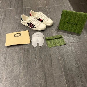 Gucci Ace vita sneakers med brodyr - Nästan helt nya Gucci Ace sneakers i vitt skinn med klassiska gröna och röda ränder på sidan samt broderad orm. Skorna är 42 men passar även 43. Baksidan har metallicröd och metallicgrön detalj med Gucci-logga. Platt sula och snörning i fram! Påse, dustbag, kyvert och skydden medföljer vid köp! Hör av er, tveka inte att skriva!😁