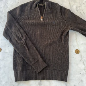 Brun stickad tröja med half zip från Morris - Säljer en brun stickad tröja från Morris i storlek M. Tröjan har ribbad struktur, lång ärm och en snygg half zip med läderdetalj vid dragkedjan. Perfekt för dig som gillar klassisk stil med en modern touch.