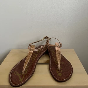 Roséguldiga sandaler från Sam Edelman - Snygga sandaler från Sam Edelman i roséguldigt syntetmaterial med reptilmönster. De har tunna remmar, platt sula och öppen tå.