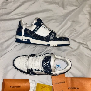 Louis Vuitton blåvita sneakers - Säljer ett par Louis Vuitton sneakers i blått och vitt med klassiskt monogrammönster. Skorna har snörning, platt sula och rund tå. Materialmix av skinn och jeans med tydliga LV-loggor på tungan och sidan. Snygga detaljer och kontrasterande färger gör dem riktigt unika.