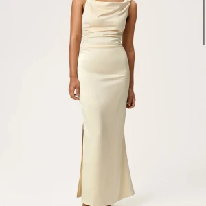 Helt oanvänd Adoore Almeria Dress med lapp kvar - Elegant långklänning i champagnefärgad satin med slits på sidan. Klänningen har en asymmetrisk axelbandslös design med en axelrem och draperad halsringning. Snygg, figurnära passform som ger en lyxig känsla till looken.