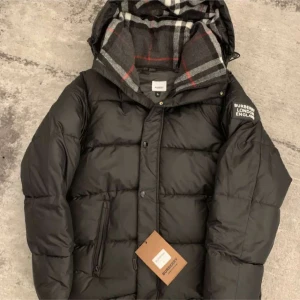 Svart pufferjacka från Burberry - Svart pufferjacka från Burberry med klassiskt rutigt foder i huvan. Jackan har dragkedja och knappar framtill, samt fickor med dragkedja. Märkeslogga på ärmen och varm, vadderad design som passar perfekt när det är kallt ute.