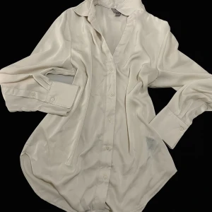 Vit glansig skjorta i silkeslook - Snygg vit skjorta med glansig finish och klassisk krage. Skjortan har långa ärmar med breda manschetter och knappar framtill. Materialet ger en lyxig silkeslook och känns mjukt mot huden. Perfekt för dig som gillar stilrena och tidlösa plagg. Storlek S men passar som M-L 