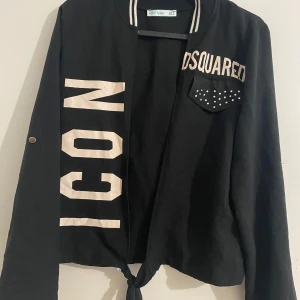 Svart jacka med tryck och rosett - Svart bomberjacka från Blue Space med stora vita tryck, dekorficka med pärlor och knytning fram. Baksidan har en cool illustration med blåvitrutig skjorta och en glittrig svart rosett. Randig krage och långa ärmar med knappdetalj.