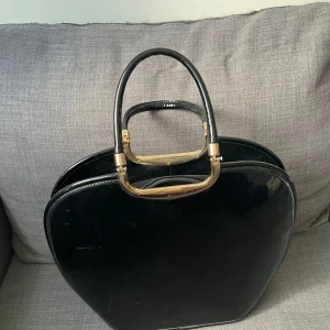 Svart handväska med gulddetaljer - Stilren svart handväska i glansigt skinn med unika guldfärgade handtag. Väskan har rundad form och dragkedja upptill. Insidan är fodrad och har flera fack för småprylar. Perfekt för dig som gillar en klassisk men ändå cool look.