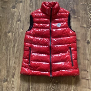Röd dunväst från Moncler med storlek 13-14 - Snygg röd dunväst från Moncler med glansig finish och svart innerfoder. Västen har hög krage, två dragkedjefickor och klassisk Moncler-logga på bröstet. Perfekt för lager-på-lager och ger en sportig vibe. Köparen betalar frakten.
